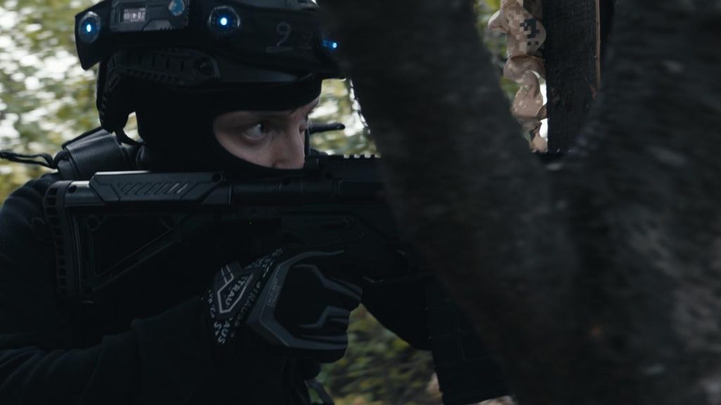 Im Wald der Outdoor-Lasertag-Arena in Wels schaut ein SWAT-Agent mit Sturmhaube und Helm durch das Zielfernrohr einer Waffe.