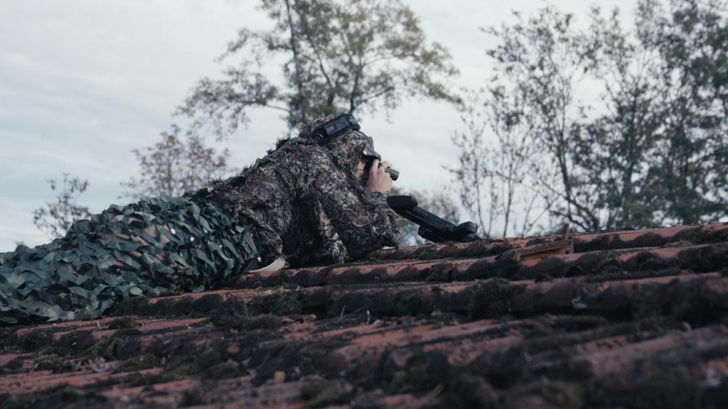 Ein Mann liegt mit dem Bauch auf einem Dach in einem Ghillie-Suit und schaut durch ein Gucker. Neben ihm liegt ein Scharfschützengewehr. Im Hintergrund ist der Wald von der Outdoor-Lasertag-Arena in Wels. Szene aus dem Projekt "The Extraction".