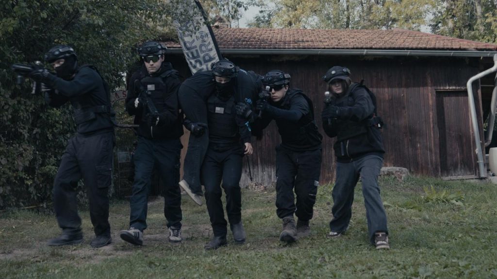 Vor einer Hütte im Wald der Outdoor-Lasertag-Arena in Wels stehen fünf SWAT-Agenten mit Helm, Sturmhaube und Sonnenbrille. Der mittlere trägt ein schwarz gekleidetes Opfer am Rücken. Sie halten schwarze Waffen in den Händen und gehen auf die Kamera zu. Szene aus dem Projekt "The Extraction" von XENTRA FILMS.