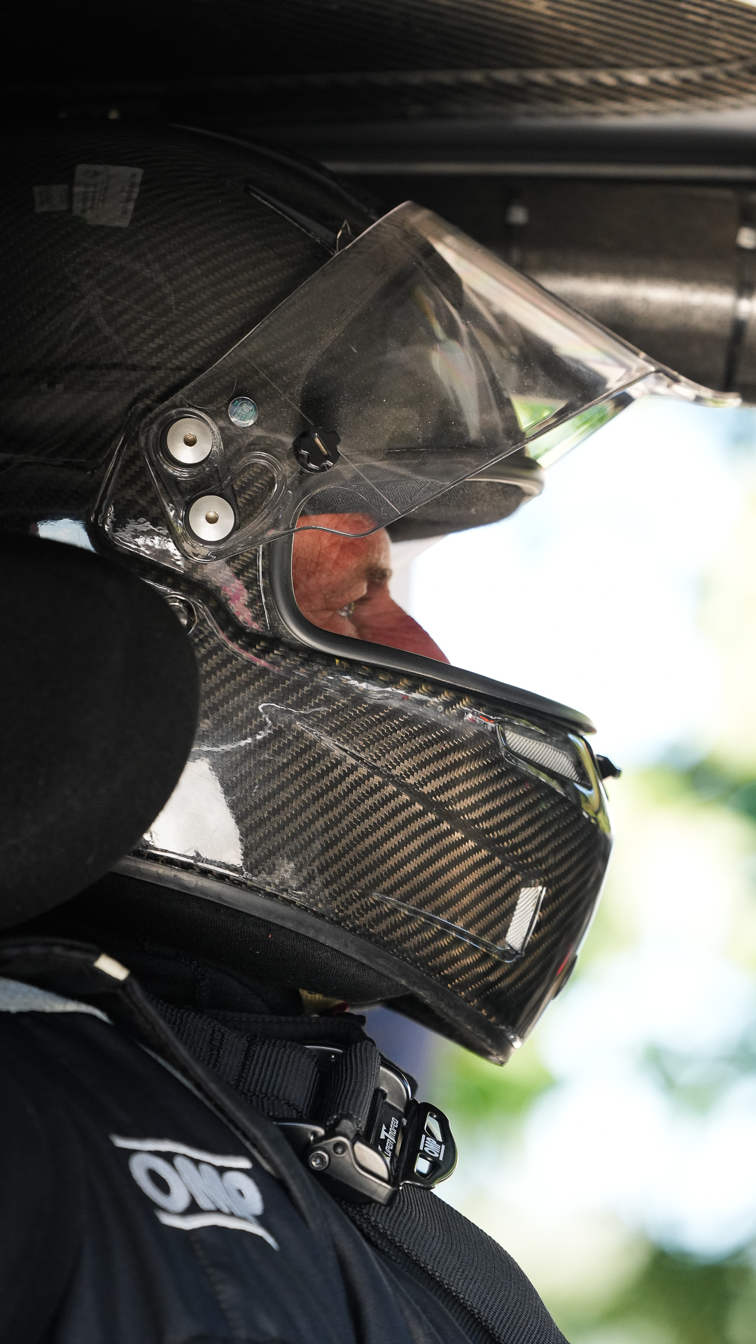 Nahaufnahme eines Carbon Helms mit geöffnetem Visier im Cockpit vor grünem Wald