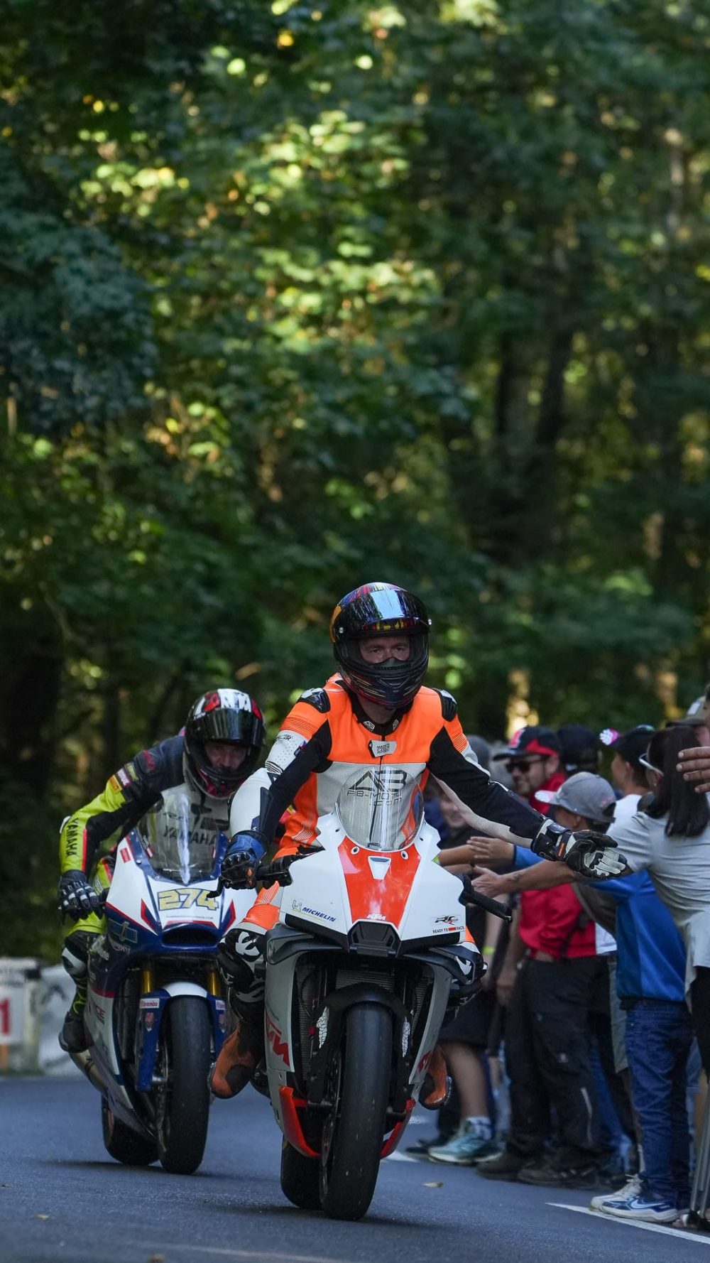Motorräder am Start Zwei Motorradfahrer in Rennkombi und Helm fahren nebeneinander auf einer Waldstraße, Zuschauer am Rand