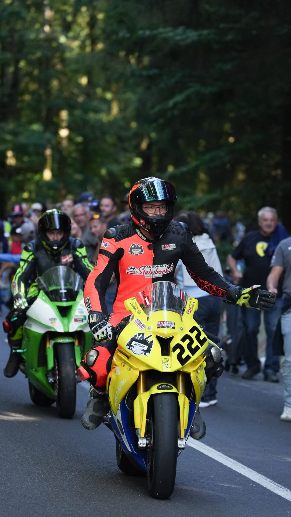 Motorräder in der Startaufstellung Motorradfahrer in rot-schwarzer Kombi auf gelbem Motorrad mit Startnummer 222, daneben ein grünes Motorrad