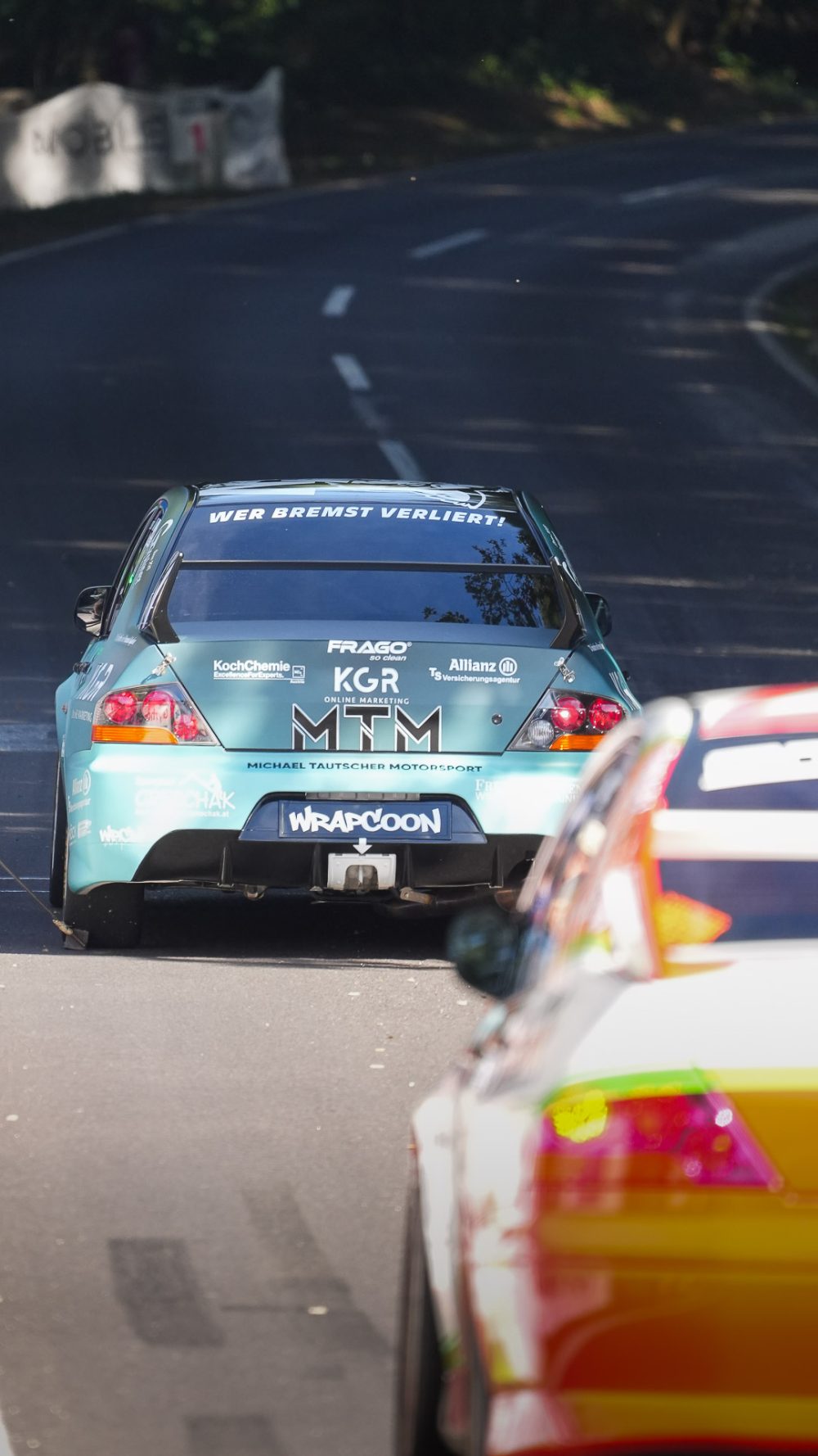 Lancer Evolution auf der Bergstraße Türkisfarbener Mitsubishi Lancer Evolution fährt auf einer kurvigen Bergstraße, im Vordergrund ein gelb-orangefarbenes Fahrzeug