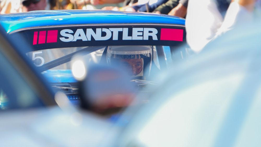 Blick durch den Vordergrund Blauer Rennwagen mit Sandtler-Schriftzug an der Windschutzscheibe, Fahrer dahinter, unscharfer Vordergrund