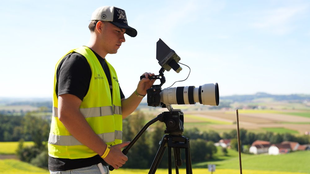 Videograf mit Stativ Kameramann in gelber Warnweste filmt mit Teleobjektiv auf Stativ vor einer Hügellandschaft