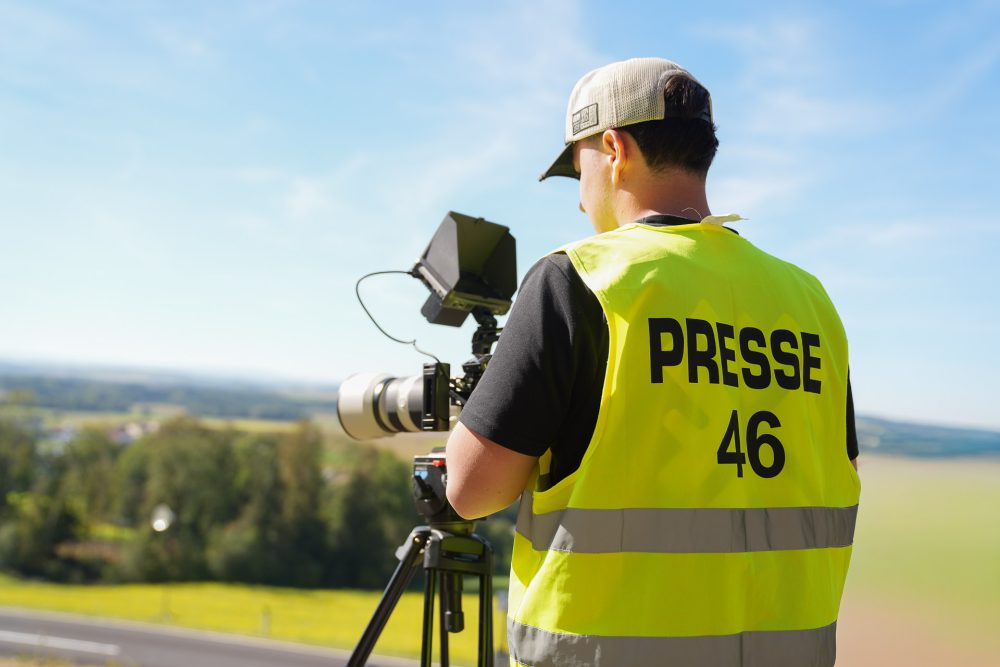 Kameramann im Einsatz Kameramann in PRESSE-Weste Nummer 46 von hinten neben Kamera auf Stativ, im Hintergrund Felder und Wald