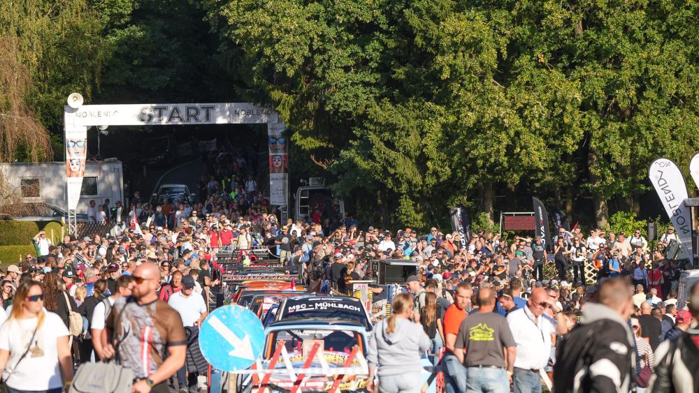 Reger Betrieb im Startbereich Menschenmenge und Rennwagen im Startbereich vor dem Startbogen zwischen hohen Bäumen