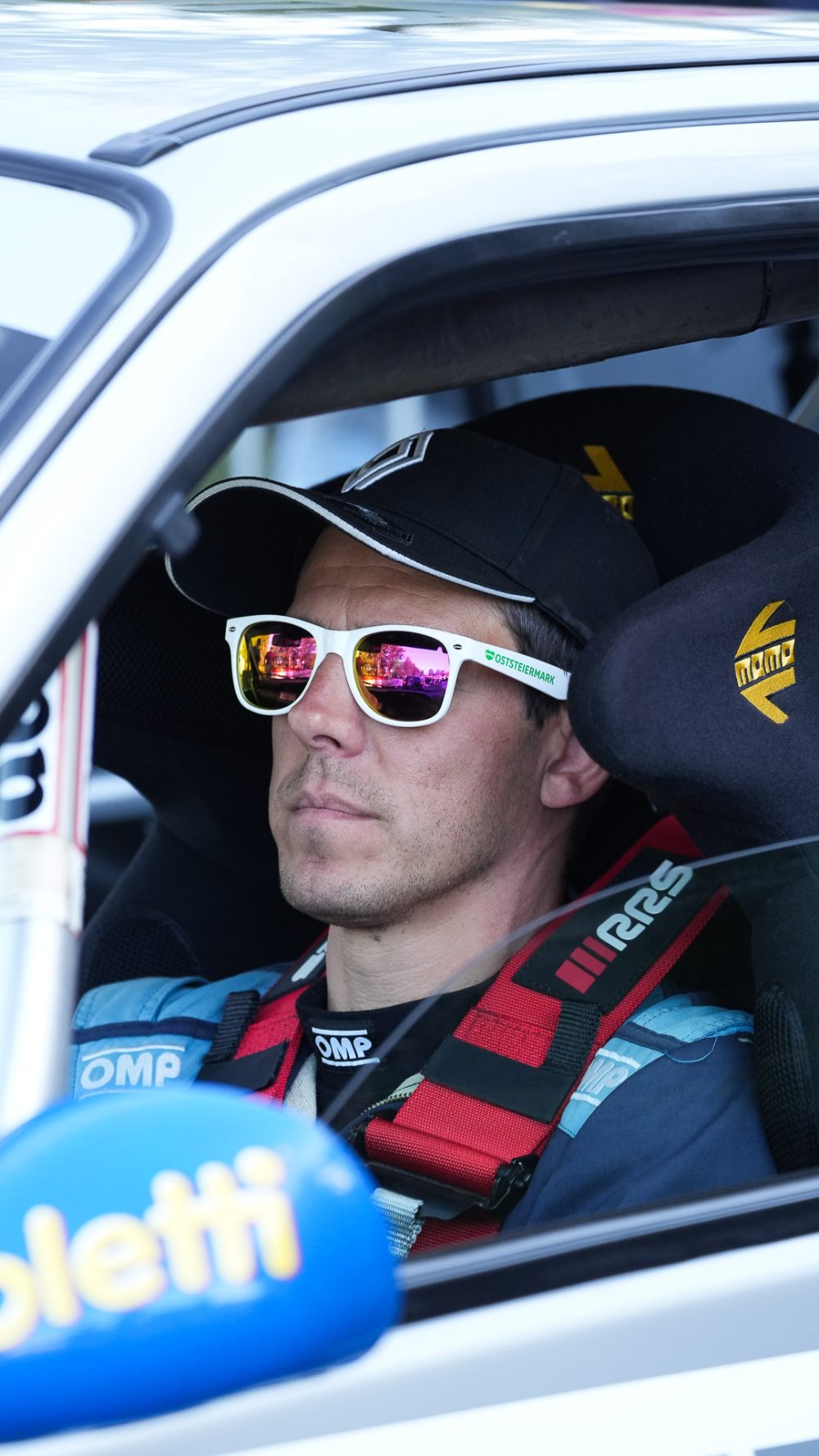 Interview im Cockpit Fahrer mit Kappe und weißer Sonnenbrille angeschnallt im Rennwagen, im Vordergrund ein blaues Mikrofon
