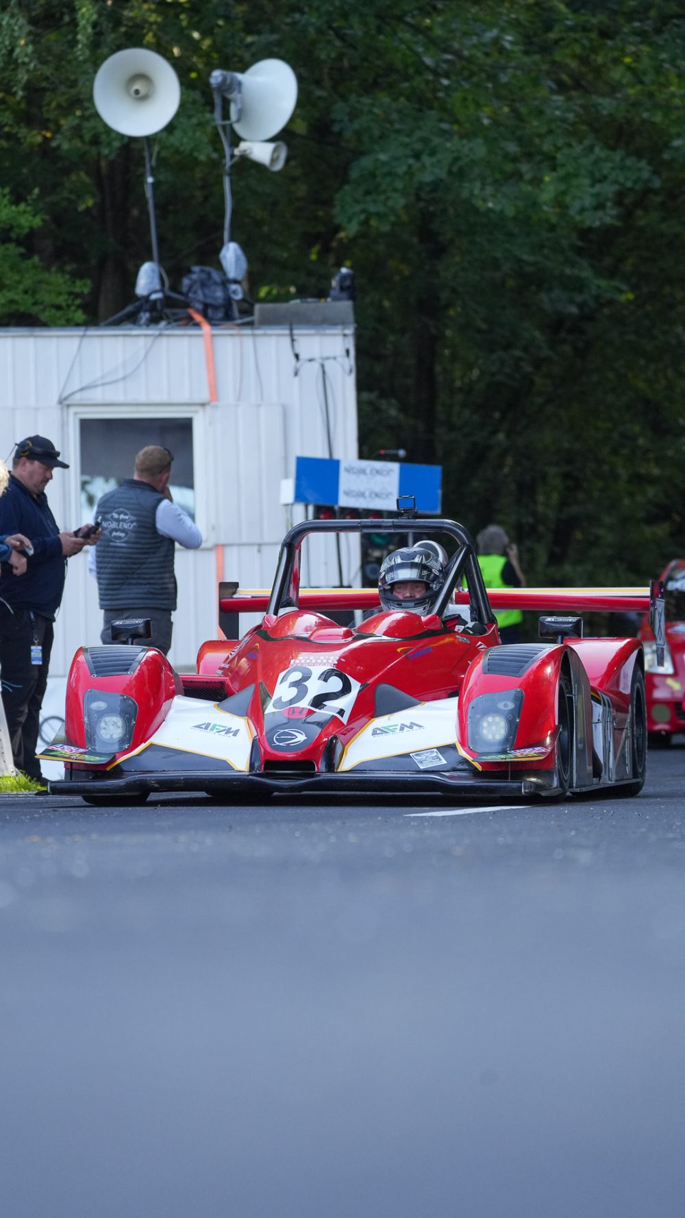 Startnummer 32 Roter Sportprototyp mit Startnummer 32 vor einem Haus mit Lautsprechern, links zwei Streckenposten