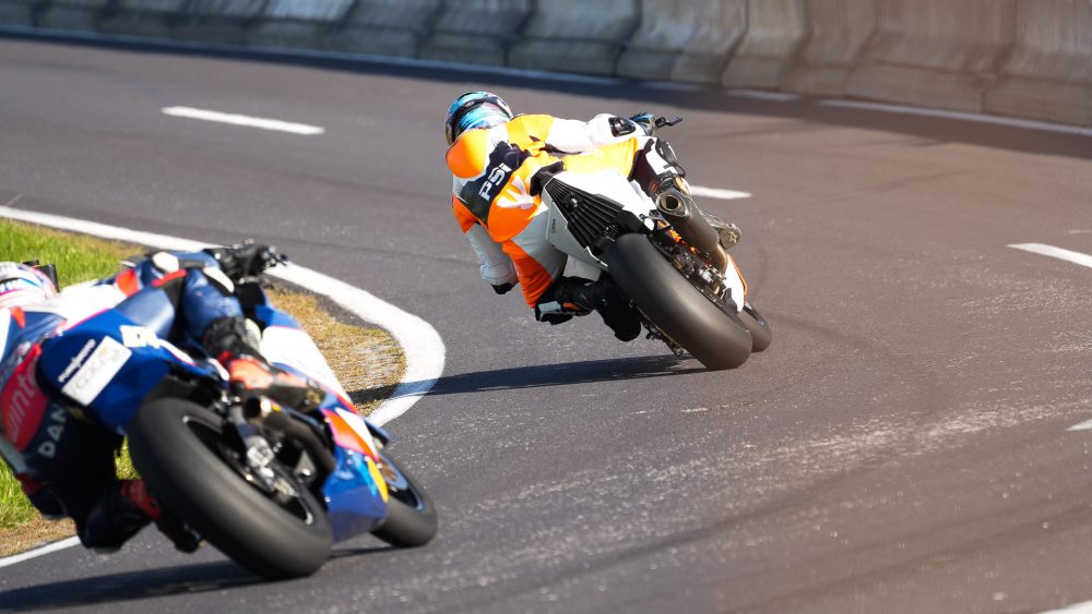 Schräglage in der Kurve Zwei Motorradfahrer in Schräglage in einer Kurve, von hinten fotografiert, neben einer Betonbarriere