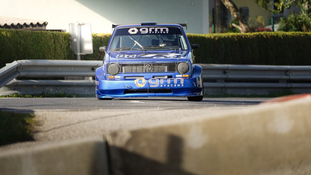 VW Golf Rennwagen Blauer VW Golf Rennwagen kommt hinter einer Leitplanke hervor, im Vordergrund ein unscharfer Betonblock
