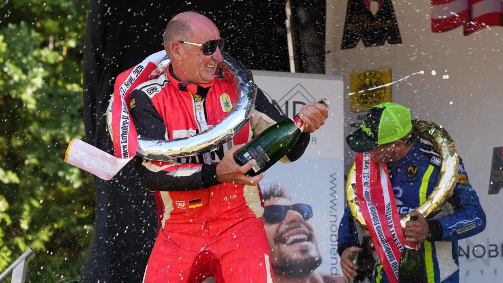 Sekt spritzt auf dem Podium Fahrer im roten Overall mit Siegerkranz und Sektflasche, dahinter ein Fahrer in Blau mit grüner Kappe
